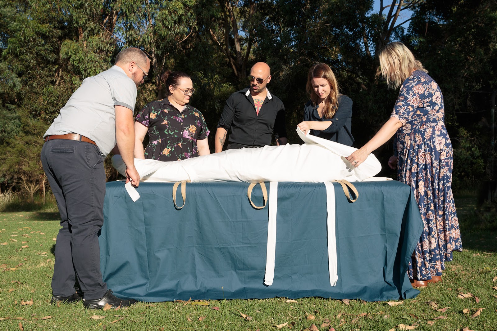 five people shrouding a body in a white shroud. set on a table covered in a teal sheet out side on the grass with trees in the back ground.